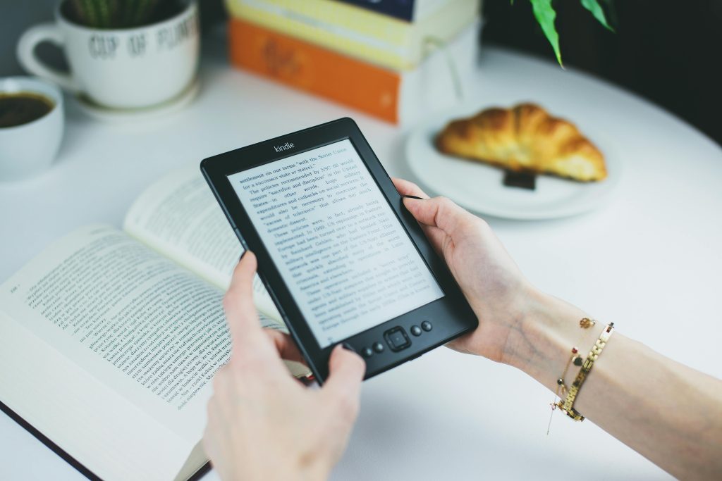 pexels-photo-76942-76942 A woman holds a Kindle while reading with a book and croissant on a table.